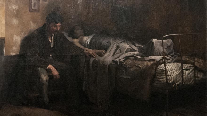 Man sitting by a bed, holding the hand of a bedridden figure in a dimly lit room